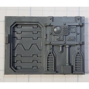May include: A gray plastic model kit piece with a detailed panel design, featuring a door with a textured surface, a control panel with buttons and a dial, and two vents with a slatted design.