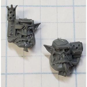 May include: Two gray plastic miniature Ork heads with detailed facial features, including sharp teeth and tusks. One head has a large, cylindrical attachment with holes, while the other has a smaller, boxy attachment. The miniatures are likely for tabletop gaming.