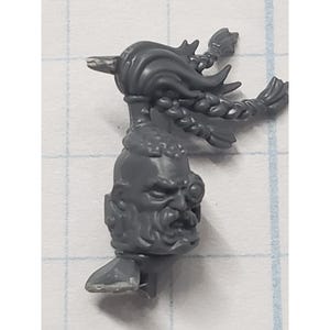 May include: A gray plastic miniature head with a stern expression, braided hair, and a beard. The miniature has a detailed design, with visible facial features and hair texture. The head is part of a larger model, likely for tabletop gaming or collecting.