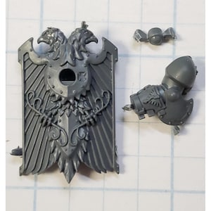 May include: A gray plastic miniature shield with a double-headed eagle design, a round hole, and ornate detailing. Also included are a shoulder pad and other small components. The pieces are likely for a tabletop game.