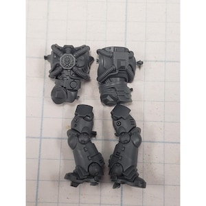 May include: Four unpainted, gray plastic miniature model components. The set includes two torso pieces with intricate mechanical detailing and two leg pieces with armored plating. The components are designed for assembly and customization.