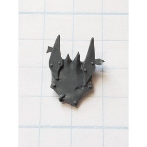 May include: A gray plastic miniature chest armor piece with a spiked design. The armor has two upward-pointing spikes and several smaller spikes along the top edge. Small rivets are visible on the surface, and there are two small arrow-like details.