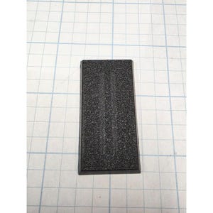 May include: A rectangular black plastic component featuring a heavily textured, granular surface finish. The object has beveled edges and a flat top profile, presented against a white grid background to provide a clear sense of its scale and shape.