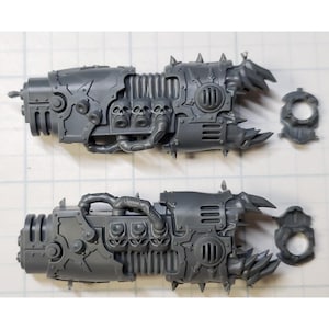 May include: Two unpainted plastic miniature weapon components. Each component features a complex, mechanical design with skulls, spikes, and a circular attachment. The pieces are gray and appear to be for a tabletop game.