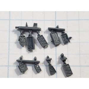 May include: A collection of small, gray plastic shields and decorative elements, likely for miniature figures or models. The shields have various geometric designs and are connected to small bars.