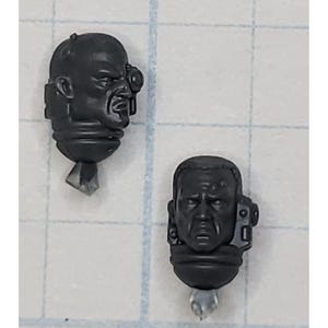 May include: Two black plastic miniature heads with detailed facial features. The heads are for use in a tabletop wargame.