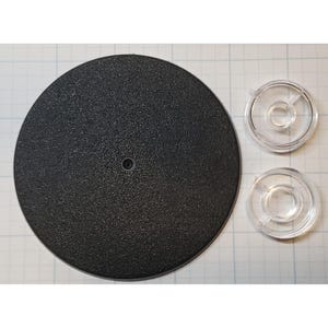 May include: A black, textured, circular base with a small hole in the center, accompanied by two clear, circular plastic pieces. The base and plastic pieces are on a white grid background.