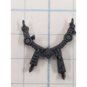 May include: A dark gray plastic miniature accessory piece featuring a curved, rope-textured bar with two circular studs, a central diamond-shaped emblem, and two dangling rectangular components with geometric engravings and pointed tips.