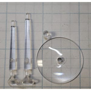 May include: Three clear plastic display stands for model figures. The stands include two tall, thin, cylindrical stands and one round base with a small center hole.