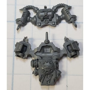 May include: Two gray plastic miniature model components. The top piece has two curved arms and a skull emblem. The bottom piece has a central body with a winged emblem and a series of rectangular shapes.