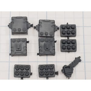May include: A collection of dark gray plastic model kit parts. The parts include rectangular blocks with circular and rectangular details, and missile-like components with multiple circular openings. The parts are arranged on a grid background.