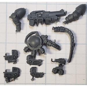 May include: A collection of gray plastic miniature model parts, including weapon components, mechanical arms, and a circular device with multiple attachments. The pieces are arranged on a grid-lined surface, suggesting a model kit or hobby project.