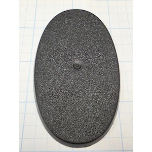 May include: An oval-shaped black plastic base with a textured, granular surface finish. The base features a single small, centered circular mounting point or peg hole. The object is positioned against a light-colored grid background.