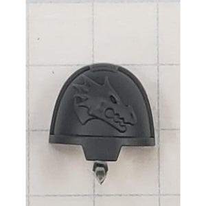 May include: A dark gray, plastic shoulder pad with a dragon head emblem. The dragon is in profile, with detailed scales and a fierce expression. The pad has a curved top and a rectangular base with a small peg.