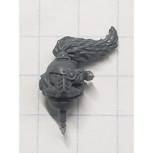 May include: A gray plastic miniature figure head with a long, flowing, textured hairpiece. The head features detailed armor elements, including a circular lens and a pointed lower section, likely for a tabletop game.