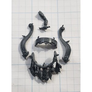 May include: A collection of gray plastic miniature figurines, including two curved serpent-like pieces, a belt, an arm holding a sword, and a decorative piece with skulls and leaves. The pieces are arranged on a grid.