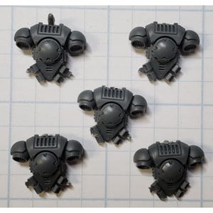 May include: Five gray plastic miniature armor pieces. Each piece features a central circular element, shoulder pads, and detailed mechanical components. The pieces are arranged on a grid background, suggesting a crafting or modeling context.