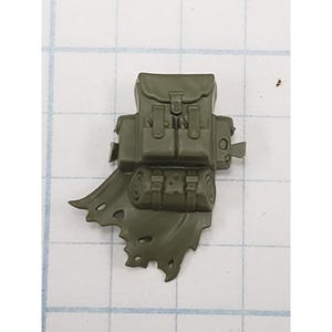 May include: An olive green miniature plastic backpack accessory featuring a main compartment with a flap, two front pockets with buckles, and a rolled sleeping bag attached to the bottom, all integrated with a tattered, flowing cape element.