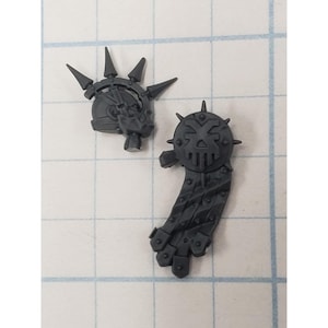 May include: Two dark gray plastic miniature components. One piece is a helmet featuring a spiked crest and a skull-like faceplate. The second piece is a shoulder pad adorned with a central skull emblem, radiating spikes, and a hanging banner.