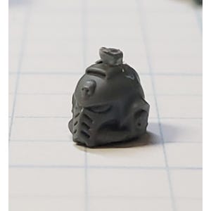 May include: A gray plastic miniature helmet with a raised detail on top. The helmet has a visor and a textured surface.
