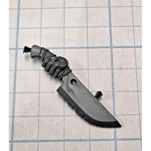 May include: A matte black miniature knife figurine featuring a handle sculpted in the shape of a clenched fist. The blade has a serrated edge, a small circular hole near the hilt, and a small protruding stud on the flat side of the blade.