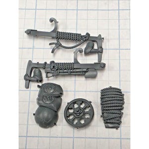 May include: A set of unpainted gray plastic miniature model components including two long pole-like weapons with rope-wrapped handles, a circular wheel-shaped piece, a rectangular piece wrapped in rope, and a curved armor plate with circular emblems.