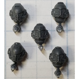 May include: Five unpainted dark gray plastic miniature helmet heads, each featuring a detailed faceplate with a grill-like mouth section, a rounded dome, and a small neck joint connector extending from the bottom of each individual helmet piece.