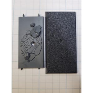 May include: Two black plastic bases for miniature figures. One base has a textured surface and the other has a raised design of a broken brick with the number "12174" on it.
