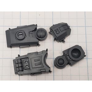 May include: Four dark gray plastic model kit components are arranged on a grid. The pieces have geometric shapes, including rectangular and circular elements. The parts appear to be from a larger model, possibly a vehicle or structure.