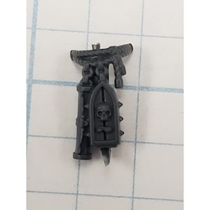 May include: A dark gray miniature accessory featuring a skull emblem on a shield, a chain, and a rope-like structure. The miniature has a spiked edge and a blade-like element at the bottom.