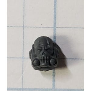 May include: A small, gray, plastic miniature helmet with a skull-like face and respirator details. The helmet has a textured surface and is likely a component of a larger model kit or figurine.