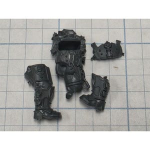 May include: A set of unpainted dark gray plastic miniature model parts including a torso piece with a chain detail, a separate chest plate featuring a skull emblem, and two distinct leg components with molded armor plating and boot details.