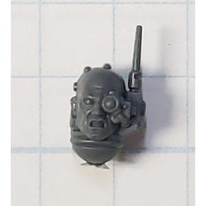 May include: A gray plastic miniature head with a single eye and a small antenna on top. The head is part of a model kit.