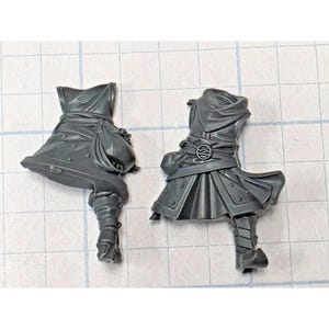 May include: Two unpainted dark gray plastic miniature torso and leg components for a tabletop game. Each piece features a sculpted tunic with a belt, a pouch, and a single armored leg. The designs include detailed fabric folds and textures.