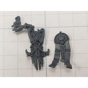 May include: Two gray plastic miniature figurines. One is a detailed figure with a skull, swords, and a banner. The other is a stylized headdress with a skull and hieroglyphs. Both are on a grid background.