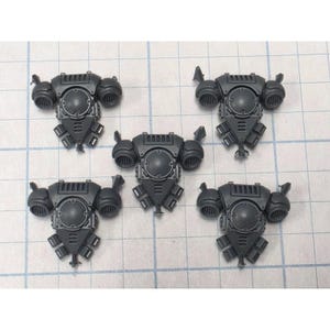 May include: Five dark gray plastic miniature backpack components, each featuring a central circular plate, two side thrusters with horizontal vents, and a triangular lower section with three small rectangular vents, arranged in a pentagonal pattern.