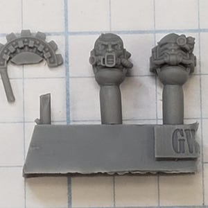 May include: Gray plastic miniature model parts. Includes two detailed helmeted heads, a cog-like piece, and a rectangular base with the letters "GW" embossed on it. The parts are for a miniature figure.
