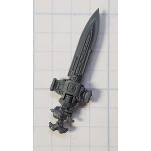 May include: A gray plastic miniature sword with intricate details and a blade with a pointed tip. The sword has a handle with a small, round, decorative piece at the end.
