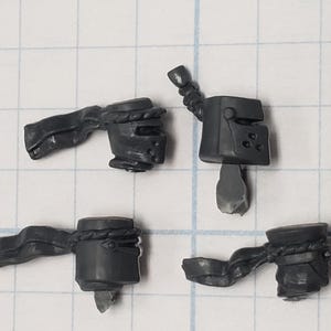 May include: Four small, dark gray plastic miniature components, possibly for a model or game. The pieces include what appear to be helmets, handles, and other structural elements. The parts are on a grid background.