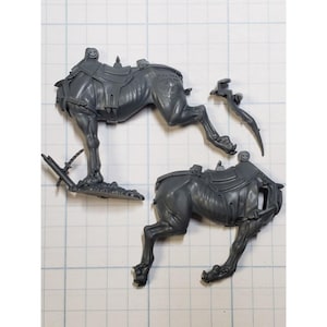 May include: Two gray plastic miniature figures of a creature with a saddle. The figures are in different poses, one is standing on its hind legs and the other is lying down.