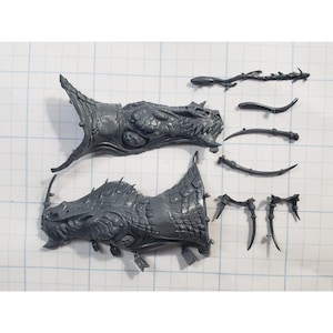 May include: Gray plastic model kit components, including two dragon heads with detailed scales and teeth, and various other parts. The pieces are arranged on a grid background, suggesting a hobby or model-making theme.