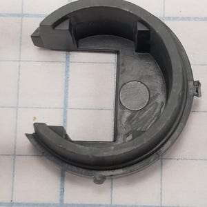 Puede incluir: Un componente de pl&aacute;stico semicircular gris oscuro con una abertura cuadrada y una hendidura circular. La pieza tiene un borde exterior redondeado y dos pesta&ntilde;as salientes. El fondo es un patr&oacute;n de cuadr&iacute;cula.