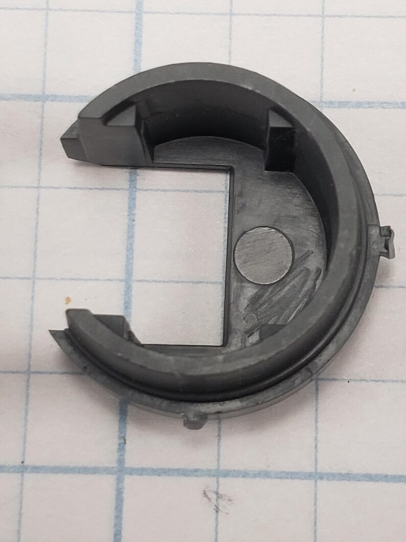 Puede incluir: Un componente de pl&aacute;stico semicircular gris oscuro con una abertura cuadrada y una hendidura circular. La pieza tiene un borde exterior redondeado y dos pesta&ntilde;as salientes. El fondo es un patr&oacute;n de cuadr&iacute;cula.
