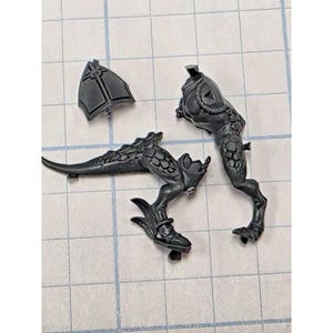 May include: Three unpainted dark gray plastic miniature model components consisting of a triangular shield piece, a torso section with textured scale-like details, and a separate leg assembly featuring a long tail and clawed feet.
