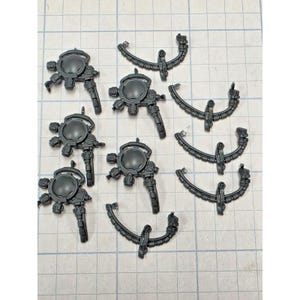 May include: A collection of dark gray plastic miniature components consisting of four circular mechanical devices with protruding nozzles and four curved, segmented structural pieces with central mounting points, arranged on a grid surface.