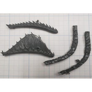May include: Four dark gray plastic model pieces, including two curved, textured sections and two spiky, ridged sections. The pieces are arranged on a grid background, suggesting they are components of a larger model or kit.