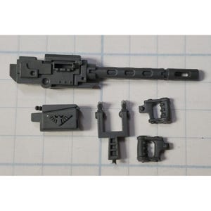 Op de afbeelding: Een verzameling ongemonteerde, grijze plastic miniature wapenonderdelen. De onderdelen omvatten een lange loop met gaten, een doos met een symbool, twee handvatten en een steunstructuur. Onderdelen voor een miniatuurmodel.