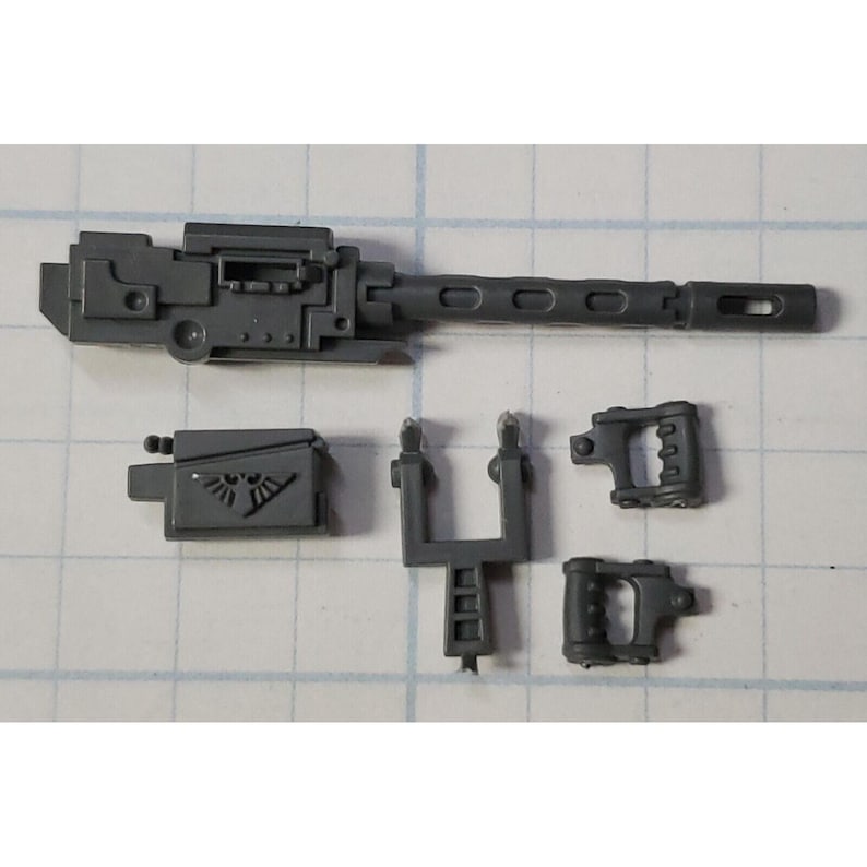 Op de afbeelding: Een verzameling ongemonteerde, grijze plastic miniature wapenonderdelen. De onderdelen omvatten een lange loop met gaten, een doos met een symbool, twee handvatten en een steunstructuur. Onderdelen voor een miniatuurmodel.