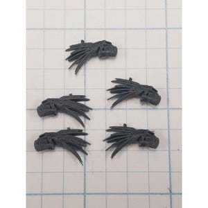 May include: Five dark gray plastic miniature creature heads, each featuring a distinct elongated snout and multiple thin, pointed crests extending backward from the skull, arranged on a grid background with one-inch square markings.
