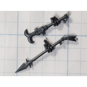 May include: Two dark gray miniature weapons, one with a hook-like end and the other with a spearhead and arrow fletching. Both have intricate details and are likely for a miniature game or model.